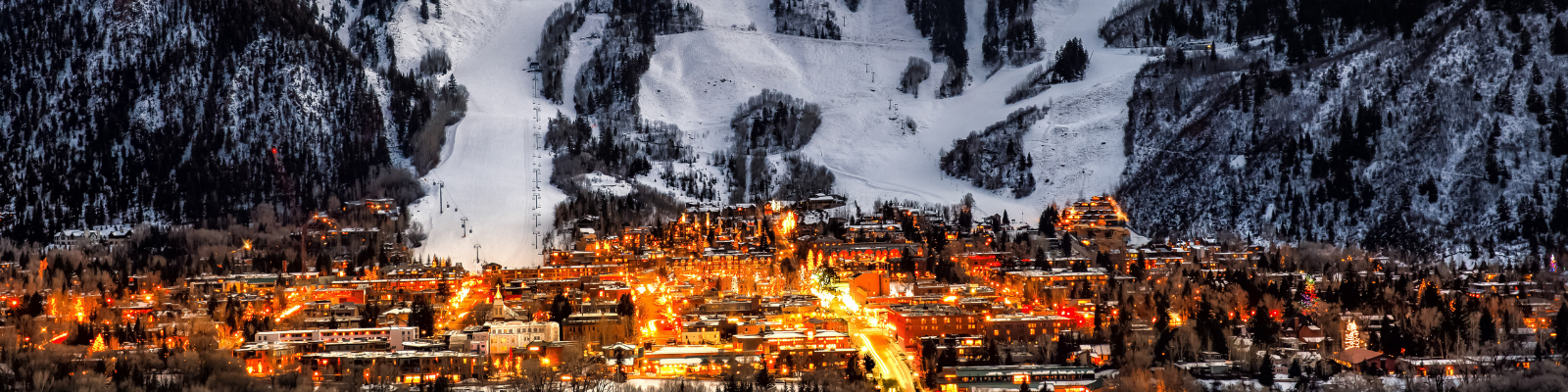 panoramic image of aspen in the evening with a row of lit up buildings in front of mountains