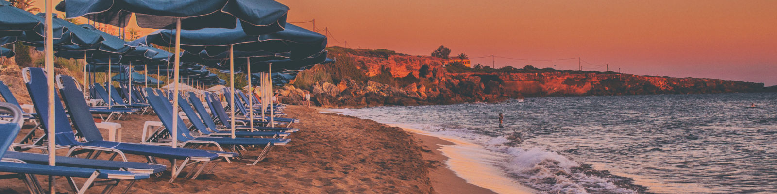 Shore of a beach at sunset with blue lounge chairs covered by blue umbrellas on the left hand side of the image and some small cliffs in the background